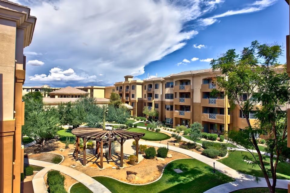 Outdoor courtyard area of a senior living facility with a wooden pergola, walking paths, green lawns, trees, and a multi-story beige building surrounding the courtyard under a partly cloudy blue sky.