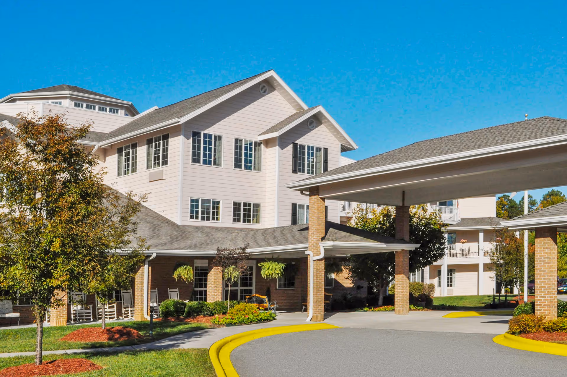 Front exterior of a light-colored multi-story senior living building with a covered drop-off porte-cochère, landscaping and trees under a clear blue sky.