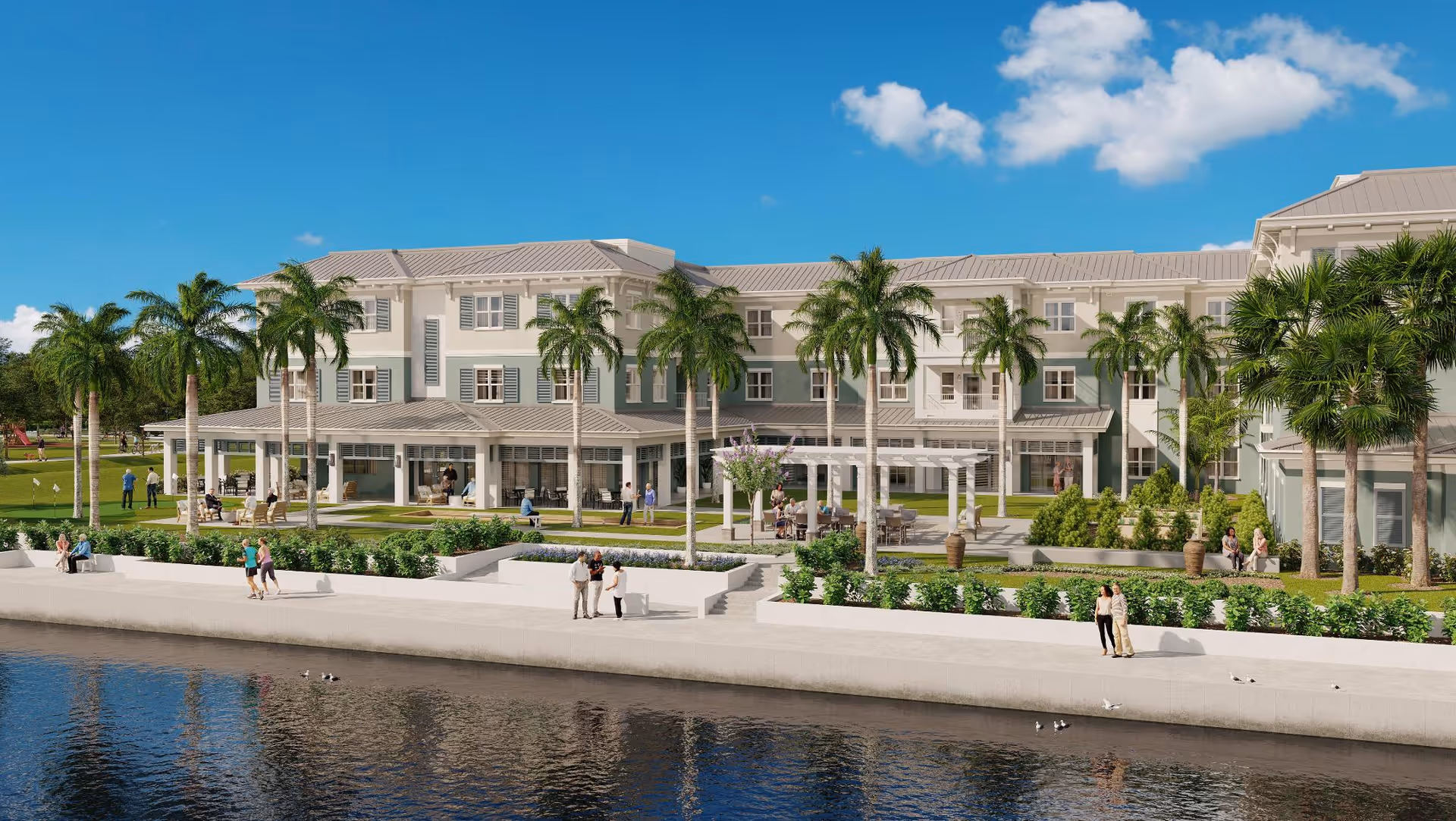 A large, modern senior living facility building with three stories, surrounded by palm trees and green landscaping. People are walking and sitting near the water's edge in front of the building, which has a clear blue sky overhead.