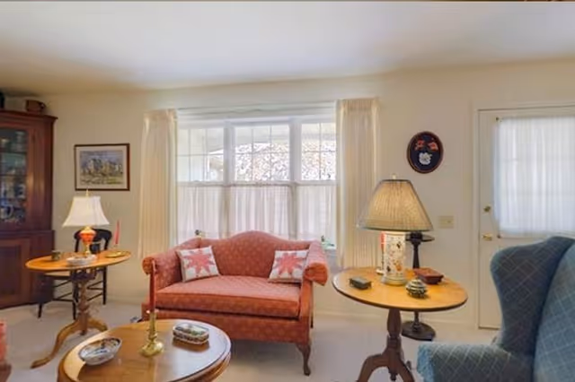 A cozy living room with a red upholstered loveseat with two patterned pillows, a blue armchair, two wooden side tables with lamps, a wooden coffee table with decorative items, a large window with sheer and cream curtains, a wooden cabinet, and framed artwork on the wall.