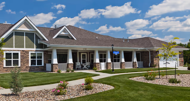 Single-story residential building with a stone and siding exterior, a covered front porch with white railings and chairs, well-maintained green lawn, landscaped flower beds with rocks and small trees, under a blue sky with scattered clouds.