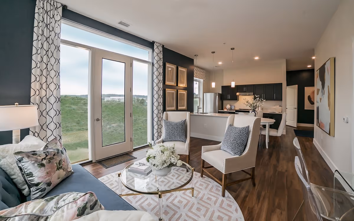 A modern open-concept living area with a dark blue sofa adorned with floral pillows, two beige armchairs with patterned cushions, and a round glass coffee table with a floral centerpiece. The room features large glass doors with patterned curtains leading outside, dark wood flooring, and a kitchen area with dark cabinets, stainless steel appliances, and pendant lighting in the background.