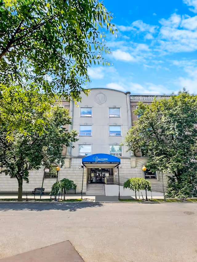 Front exterior view of a multi-story senior living facility named Belvedere Senior Living, with a blue awning above the entrance, surrounded by green trees and a clear blue sky.