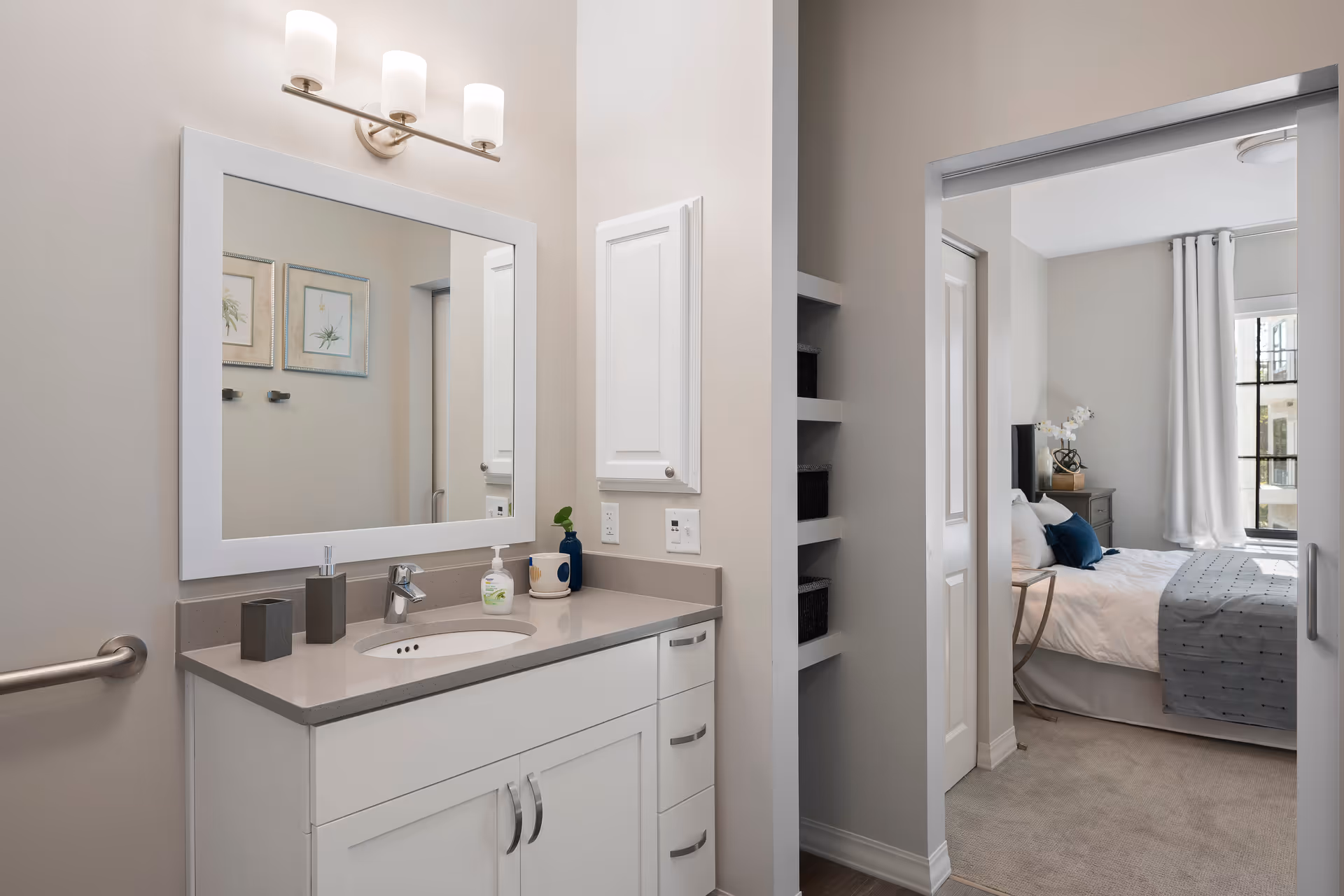 A clean and modern bathroom vanity with a large rectangular mirror above it, three light fixtures mounted on the wall, and various toiletries on the countertop. To the right, an open doorway leads to a bedroom with a neatly made bed, white curtains, and a window letting in natural light.
