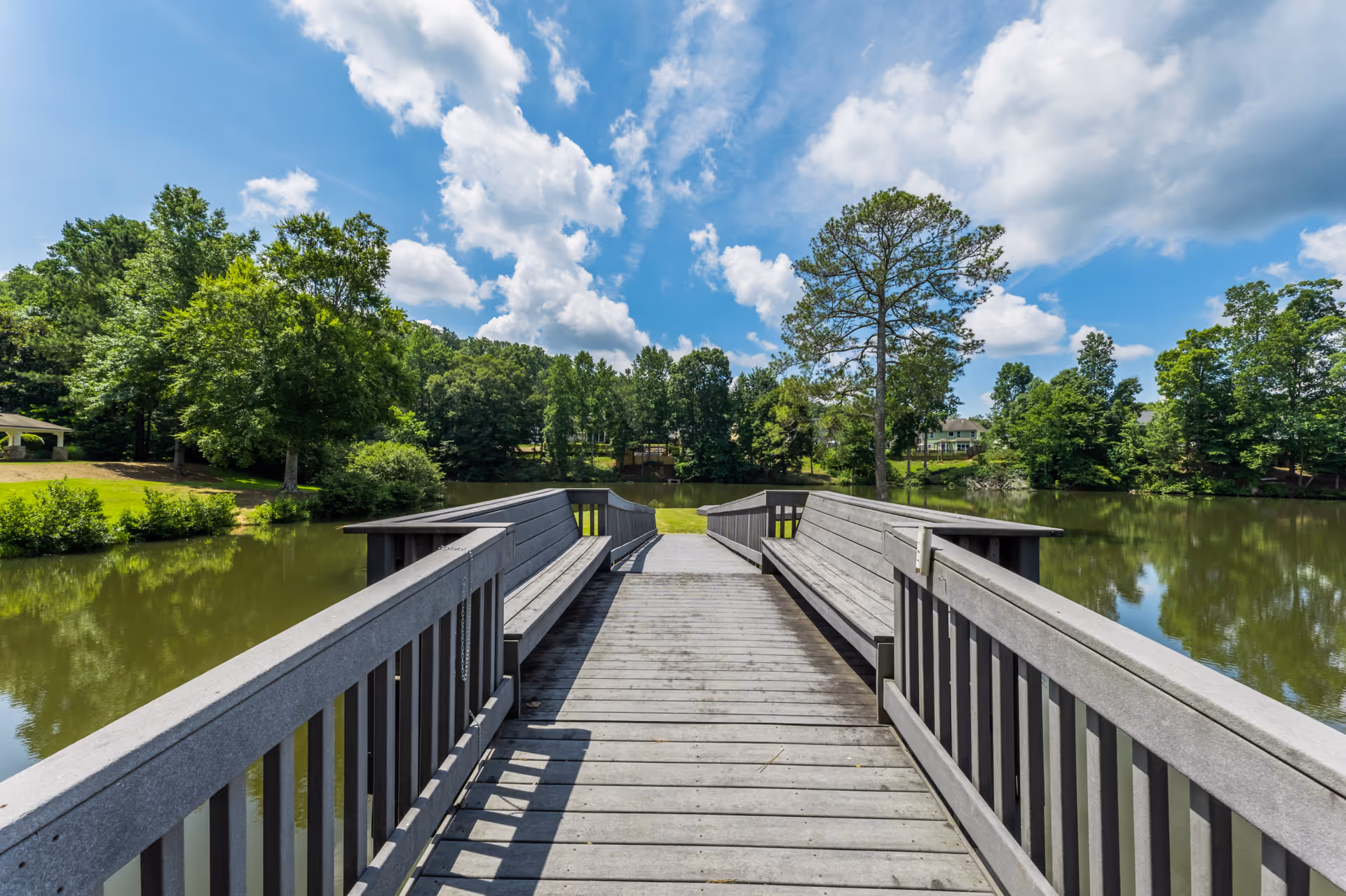 A wooden dock with built-in benches on both sides extends over a calm lake surrounded by lush green trees under a partly cloudy blue sky.