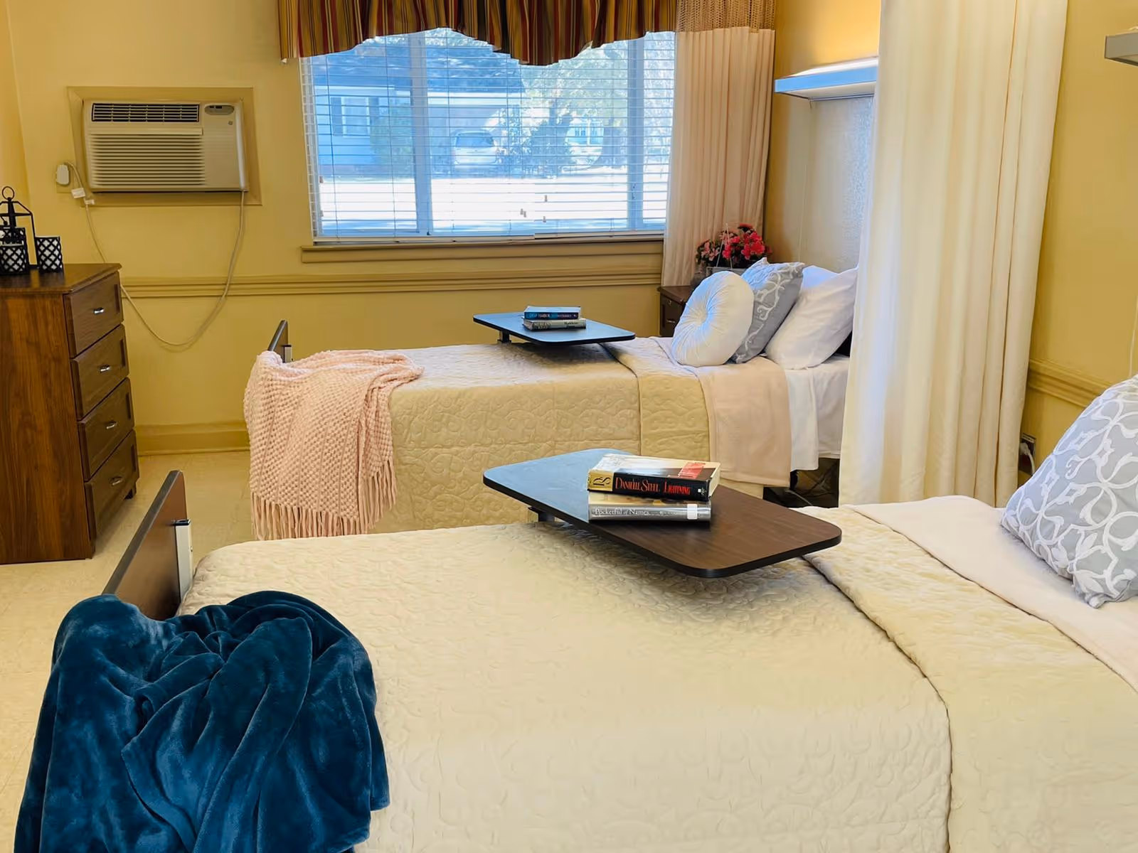 A bright senior care bedroom with two twin beds, overbed tables holding books, a dresser, and a window air conditioner.
