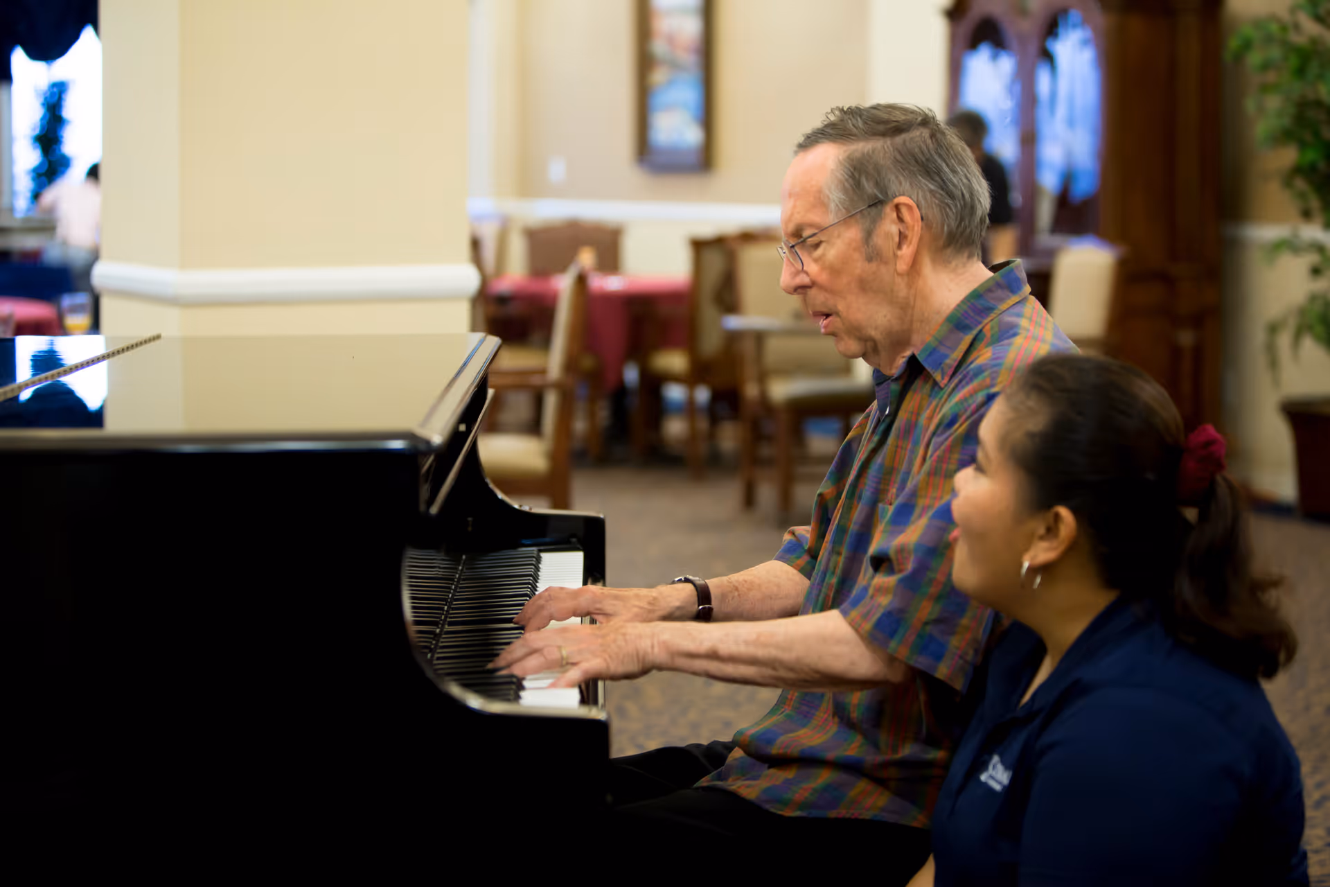 An elderly man playing a black grand piano in a common area of a senior living facility, with a woman sitting beside him watching and smiling. The background shows tables and chairs in a softly lit room.