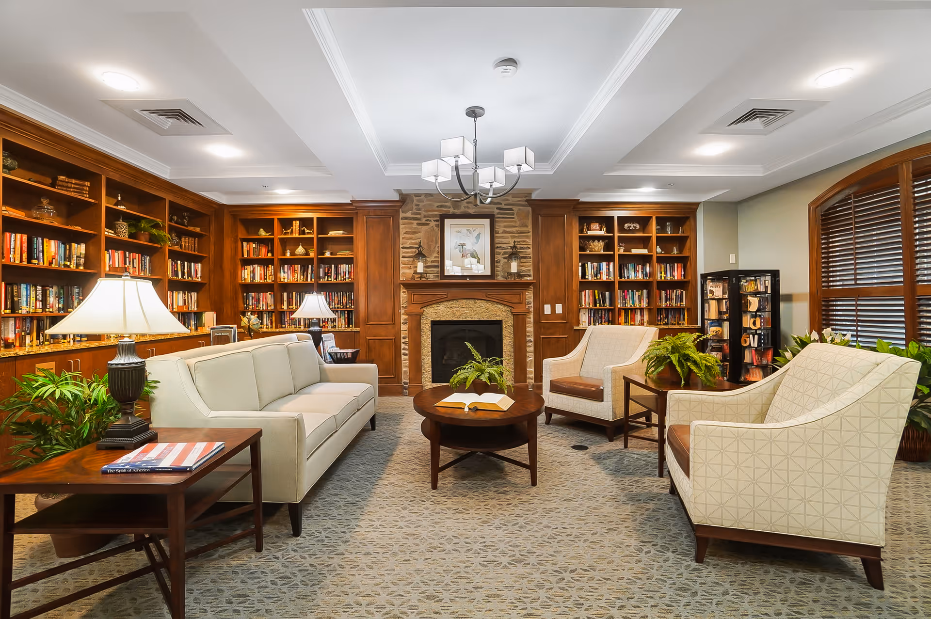 A cozy living room with a beige sofa, two armchairs, a round wooden coffee table with an open book, and side tables with lamps and plants. The room features built-in wooden bookshelves filled with books, a stone fireplace with a framed picture above it, and a large window with wooden blinds.