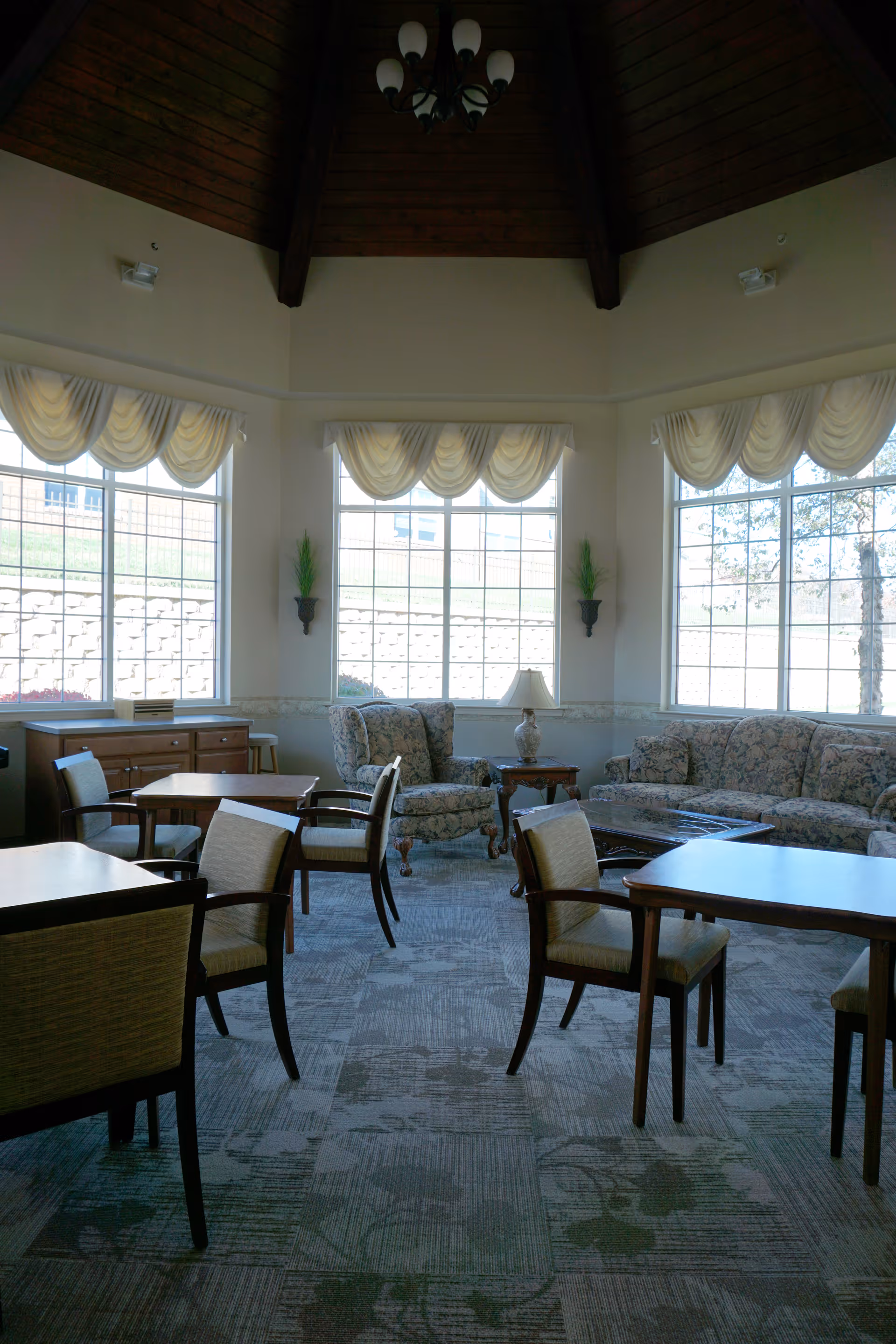 Sunlit communal lounge with tables and chairs, upholstered sofas and armchairs beneath a high wood-paneled ceiling.