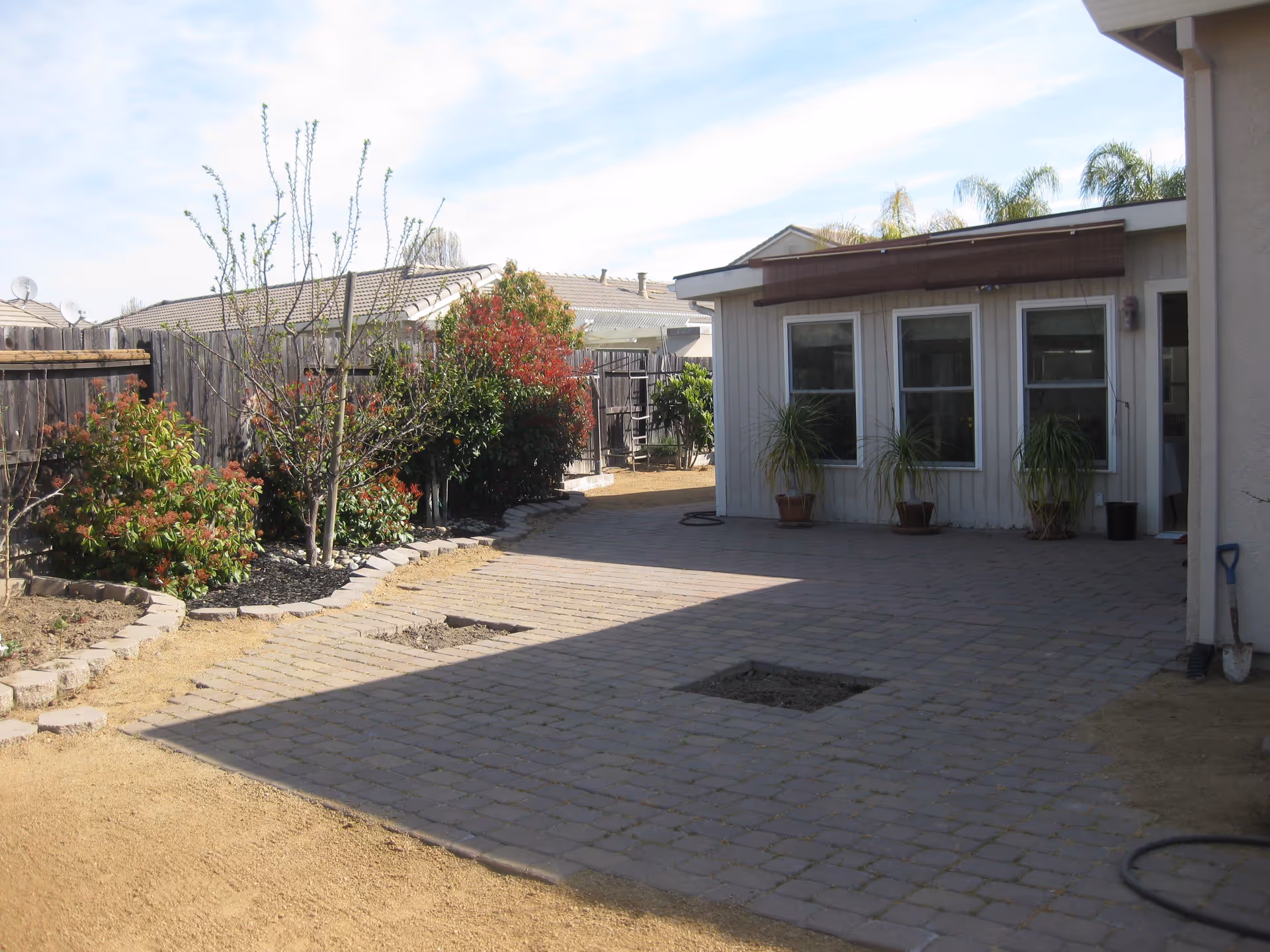 Outdoor patio area with paved stone flooring and a small square patch of soil in the center. There are three potted plants placed against the wall of a light-colored building with three windows. To the left, there is a garden bed with various shrubs and small trees, bordered by a wooden fence. The sky is partly cloudy.