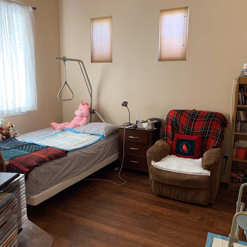 A senior bedroom with a single bed fitted with a patient lift/trapeze, a bedside table with lamp and items, and a plaid-covered recliner chair.