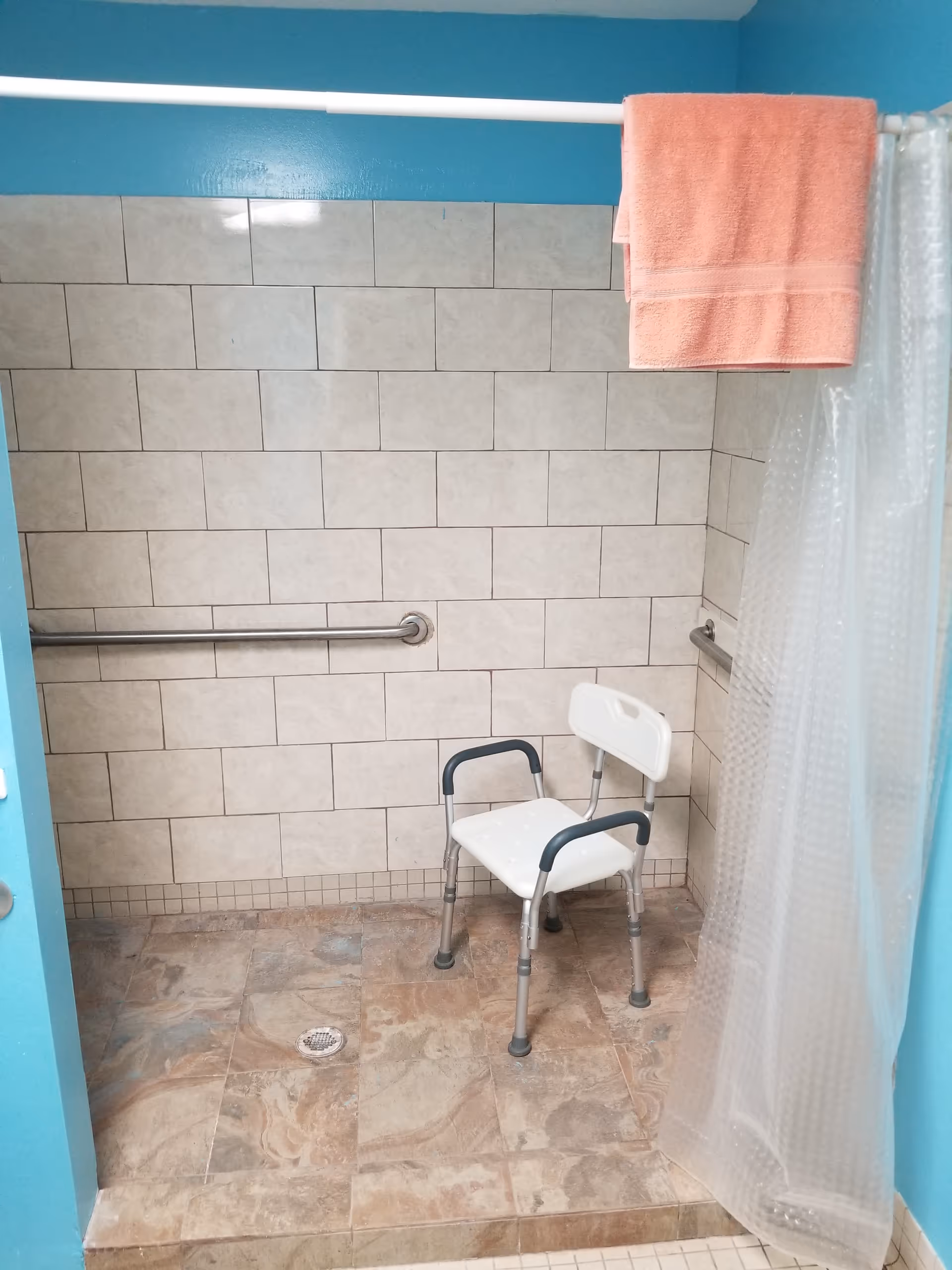 A tiled shower area with beige wall tiles and a marbled floor. There is a white shower chair with armrests and a backrest placed inside the shower. A metal grab bar is mounted on the wall for support. A clear plastic shower curtain is partially drawn to the side, and a peach-colored towel hangs over the shower curtain rod. The walls outside the tiled area are painted blue.