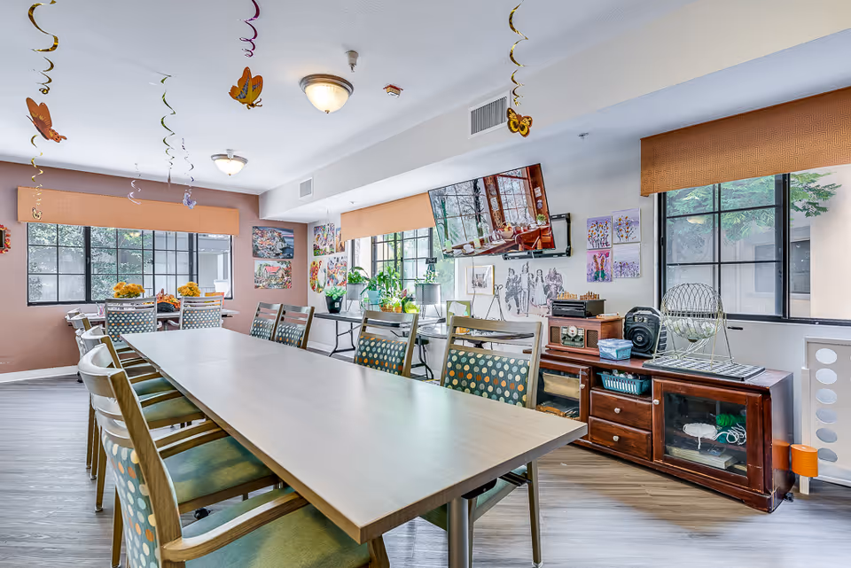 A bright and spacious activity room in a senior living facility with a long table surrounded by chairs featuring polka dot upholstery. The room has large windows with orange valances, colorful butterfly decorations hanging from the ceiling, and various artworks on the walls. There is a TV mounted on the wall, a wooden cabinet with a bingo cage and other game equipment, and several potted plants on a side table.