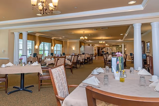 A spacious dining room with multiple tables covered in beige tablecloths, each set with white napkins, glasses, and cups. The room features large windows with yellow valances, white columns, and warm lighting from chandeliers and wall sconces. Chairs with patterned upholstery surround the tables, and a menu or informational stand is visible on one table.