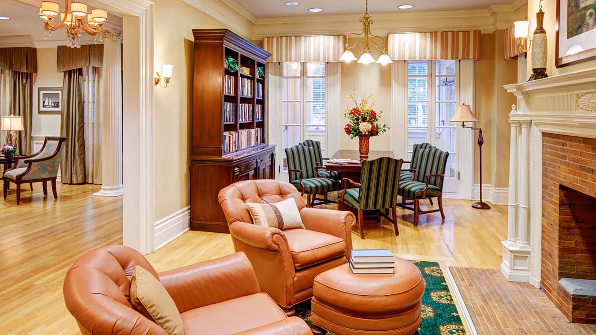 A cozy living room area with two tan leather armchairs and a matching ottoman, a green patterned rug, a wooden bookshelf filled with books, a dining table with four striped chairs, a fireplace with a white mantel, and large windows with striped valances letting in natural light.