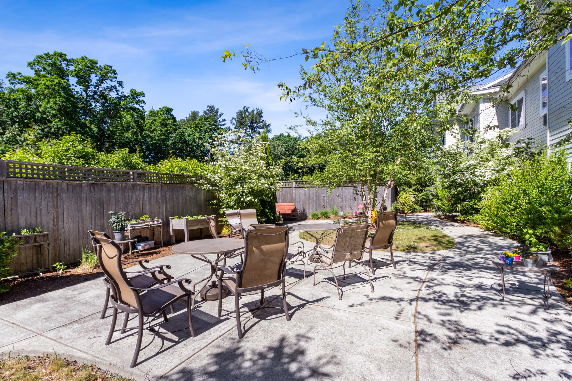 Outdoor patio area at The Enclave of Franklin with metal chairs and tables arranged on a concrete surface, surrounded by greenery, trees, and a wooden fence under a clear blue sky.
