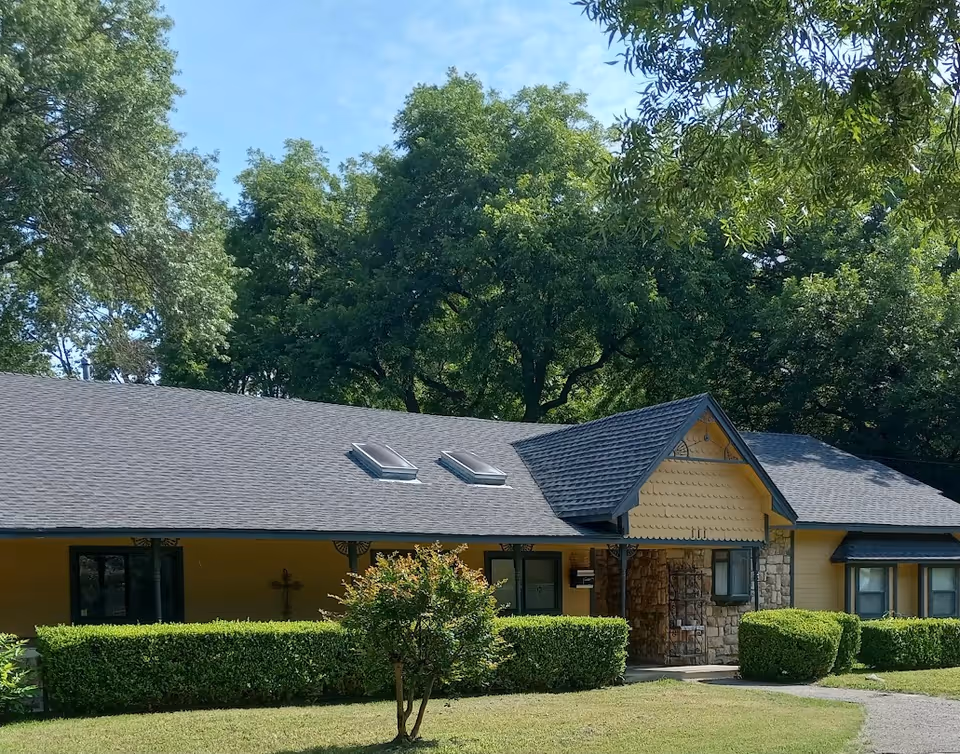 Exterior view of a single-story assisted living facility building with a gray shingled roof, yellow siding, and stone accents. The building is surrounded by green bushes, a small tree, and large leafy trees in the background under a partly cloudy sky.