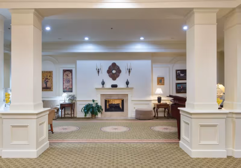 A spacious, well-lit living room area with a central fireplace decorated with a large flower-shaped wall clock and two wall sconces. The room features beige walls with white trim, two large columns, framed artwork on the walls, a green potted plant near the fireplace, a table with a lamp, and comfortable seating including chairs and an ottoman. The carpet has a patterned design with circular motifs.