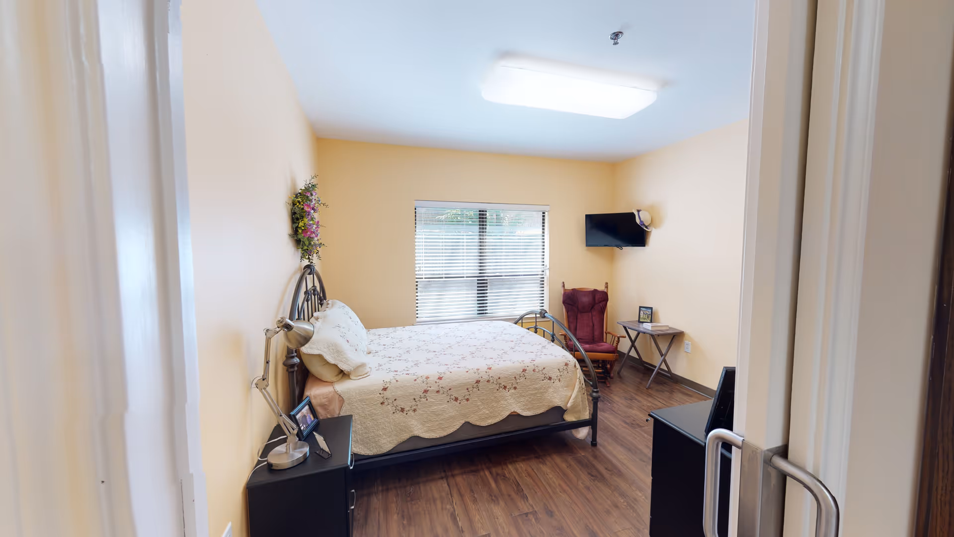A cozy bedroom with light yellow walls and wood flooring. The room features a metal-framed bed with a floral quilt, a bedside table with a lamp and picture frame, a window with blinds, a wall-mounted TV, a red cushioned chair, and a small folding table with a framed photo on it.