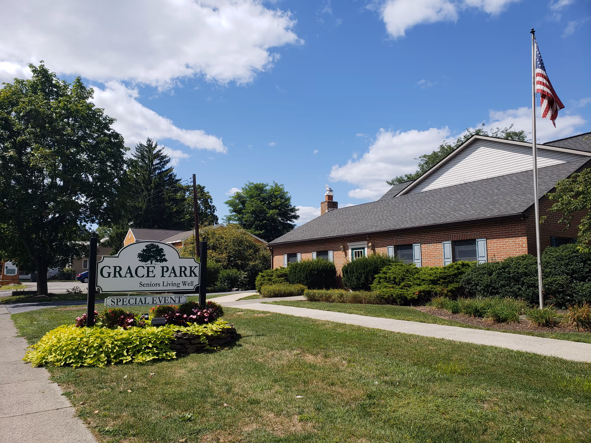 Exterior front view of Grace Park Senior Living with its entrance sign, landscaped flowerbed, walkway, and an American flag.