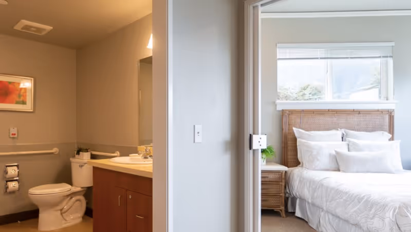 View of a bedroom and bathroom in a senior living facility. The bedroom features a bed with white bedding, a wicker headboard, a nightstand with a plant, and a window with blinds. The bathroom includes a toilet, a sink with a countertop, a mirror, and grab bars on the walls.