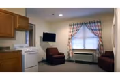 Small studio-style living area with a kitchenette on the left, two upholstered chairs facing a wall-mounted TV, and a window with plaid curtains above an air conditioning unit.