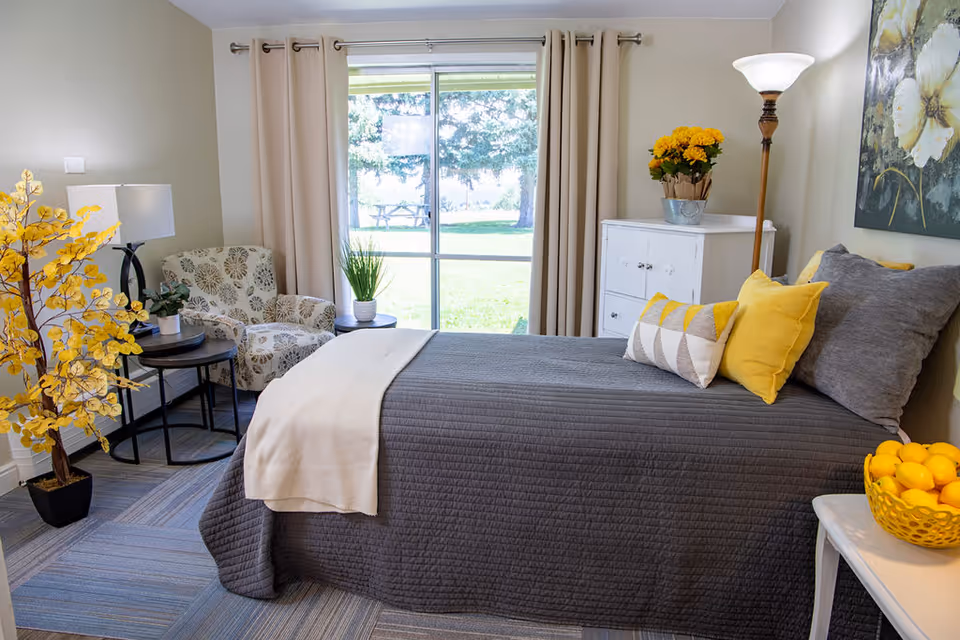 A bright bedroom with a single bed dressed in gray bedding and yellow accent pillows, an armchair, side tables, plants, and a large window looking out to a lawn.