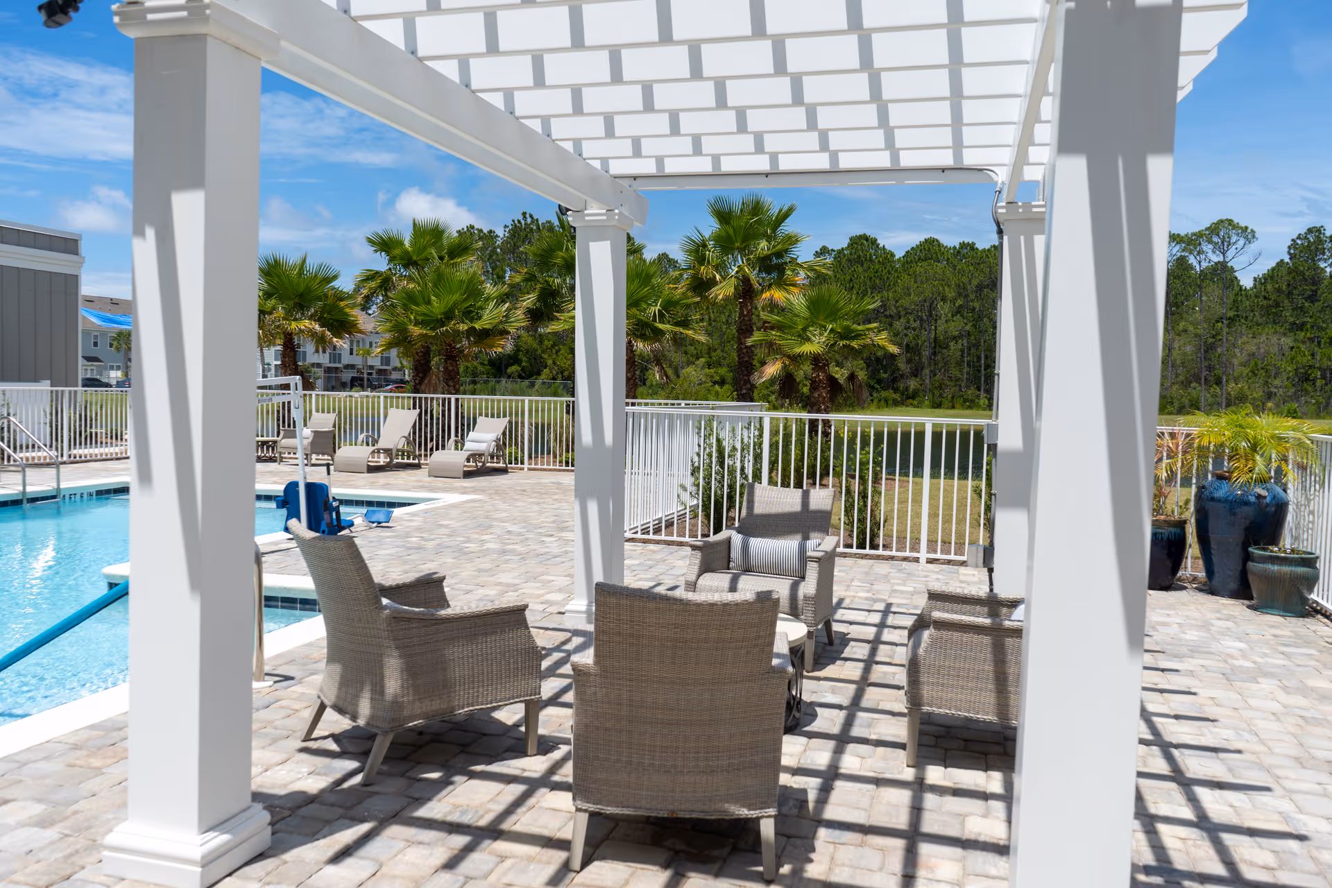 Outdoor seating area under a white pergola with four wicker chairs arranged around a small table on a paved patio next to a swimming pool. Palm trees and greenery are visible in the background under a blue sky.