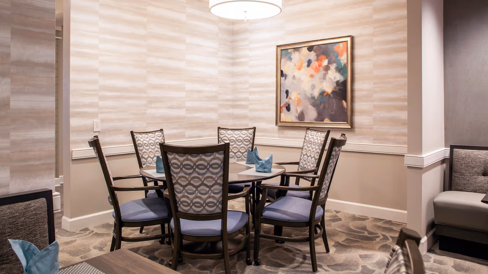 Round dining table with six patterned chairs, folded napkins, and abstract artwork on the wall in a softly lit dining room.