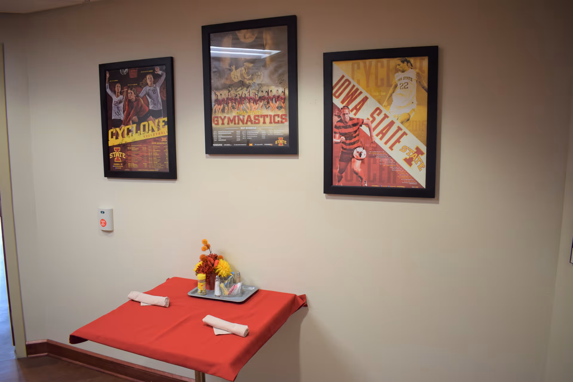 A small table covered with a red tablecloth set against a beige wall. On the table are two rolled napkins and a tray with condiments and a small flower arrangement. Above the table, three framed sports posters related to Iowa State University are hung on the wall.