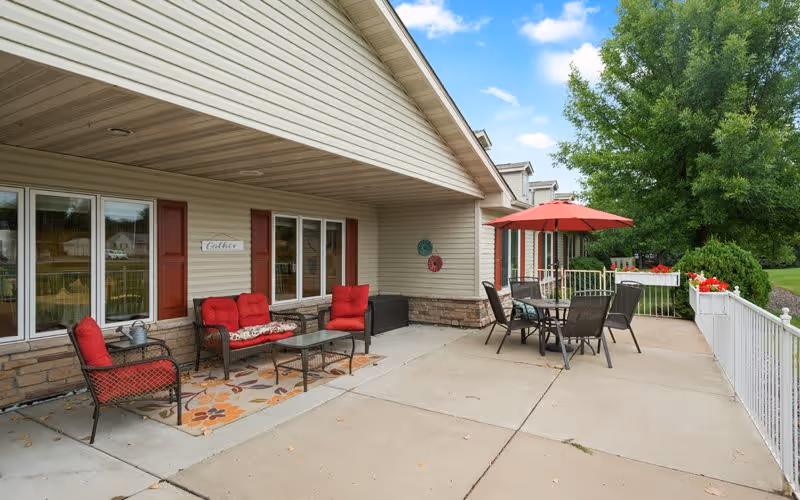 Outdoor patio area at Encore Assisted Living and Memory Care at Hugo with seating arrangements including cushioned chairs and a loveseat with red cushions, a glass coffee table on a floral rug, and a dining table with chairs under a red umbrella. The patio is adjacent to a beige building with windows and red shutters, surrounded by greenery and a white railing with flower boxes.