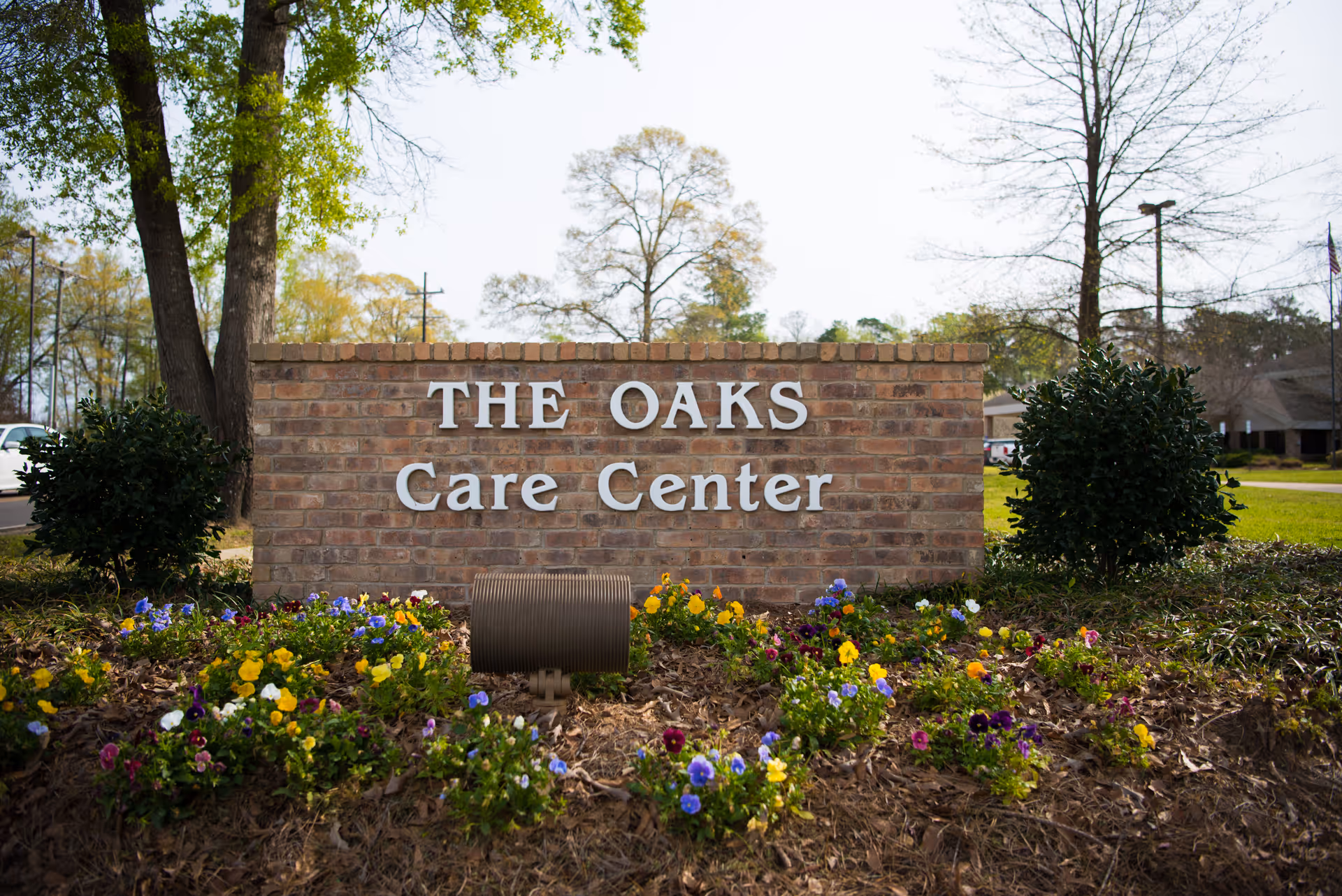 A brick sign with white letters that reads 'THE OAKS Care Center' surrounded by colorful flowers and greenery, with trees and a building visible in the background.