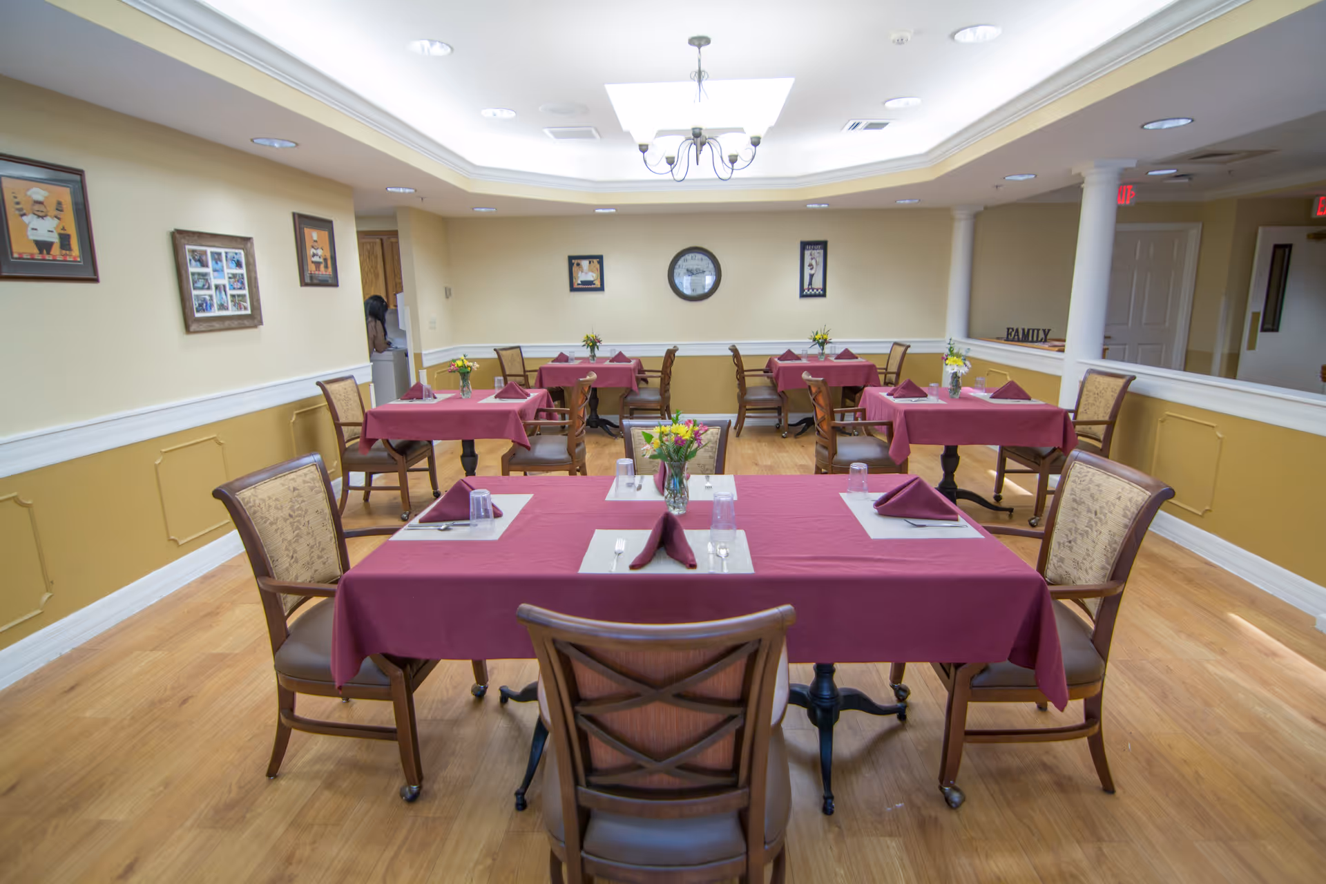 Dining room with multiple tables set with burgundy tablecloths, chairs, and floral centerpieces under a ceiling light.
