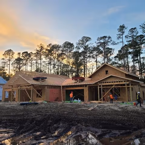 Wood-framed building under construction with workers on site and pine trees and a sunset sky in the background.