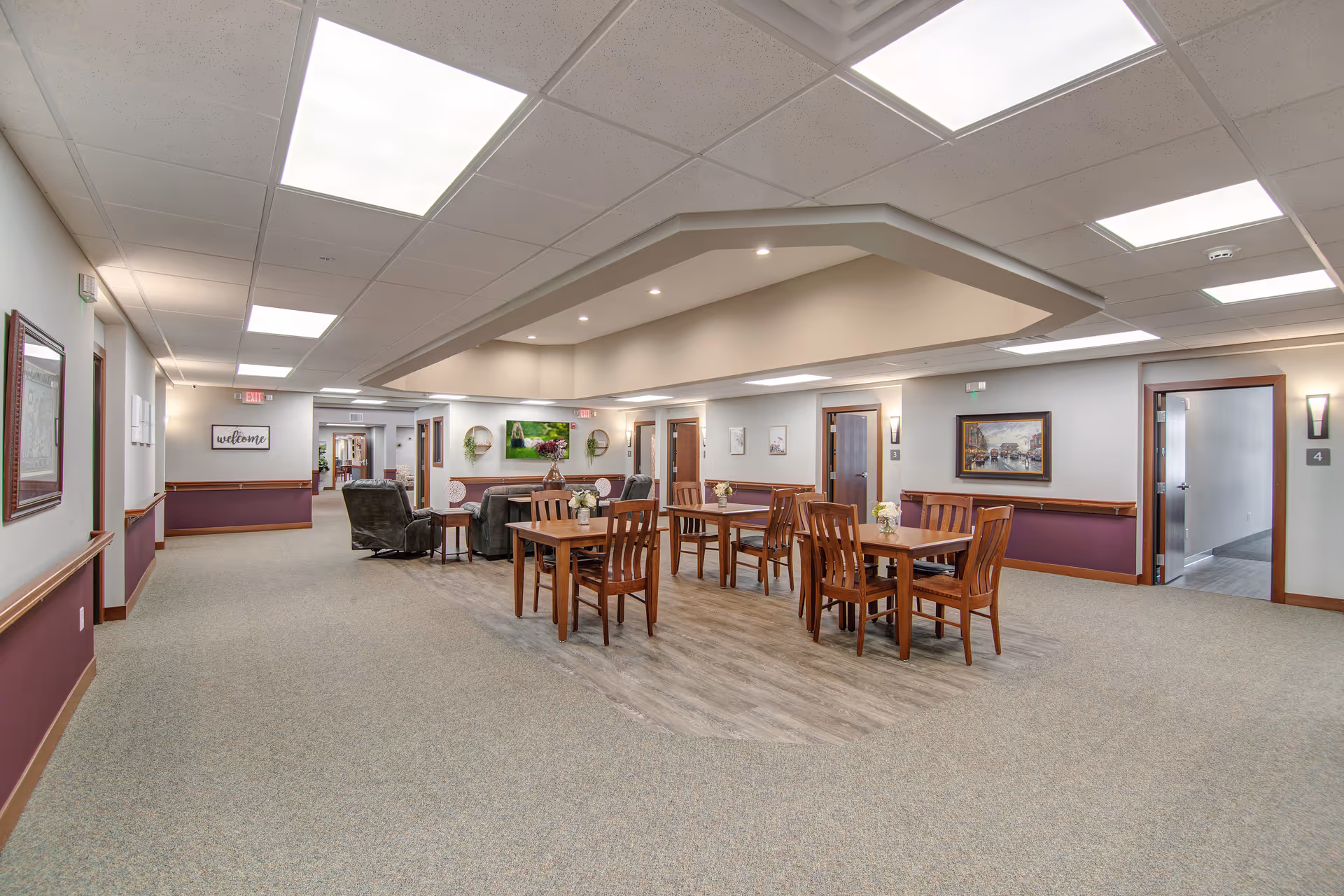 A spacious and well-lit common area in a senior living facility featuring several wooden tables with chairs arranged on a wood-floored section. Comfortable armchairs and a sofa are placed near the back wall, which is decorated with framed artwork and plants. The surrounding area has carpeted flooring and multiple doorways leading to other rooms. The ceiling has recessed lighting and large fluorescent panels.