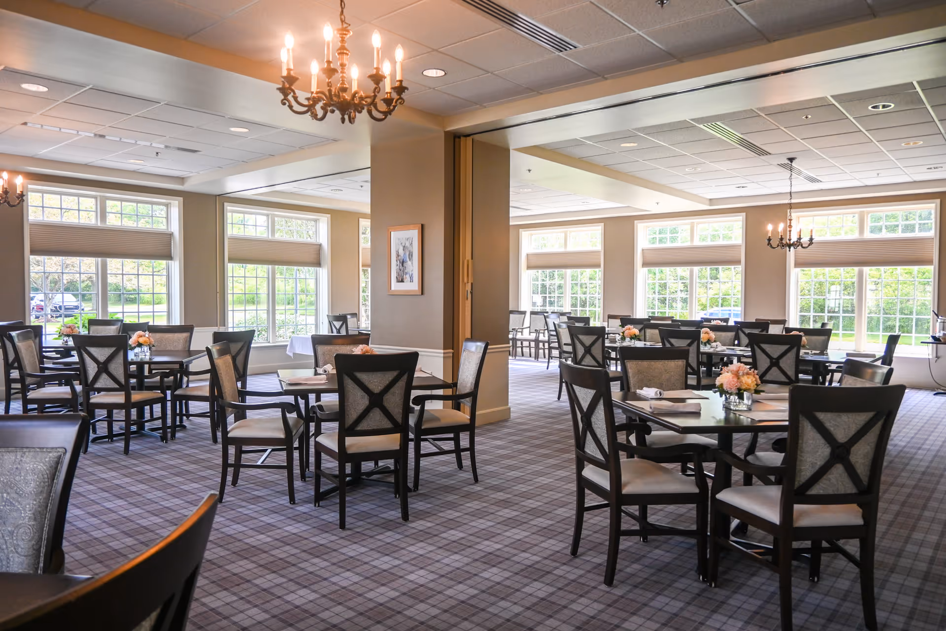 A bright and spacious dining room with multiple tables and chairs arranged neatly. Each table has a small floral centerpiece. Large windows allow natural light to fill the room, and chandeliers hang from the ceiling providing additional lighting. The carpet has a plaid pattern, and the walls are painted in neutral tones.