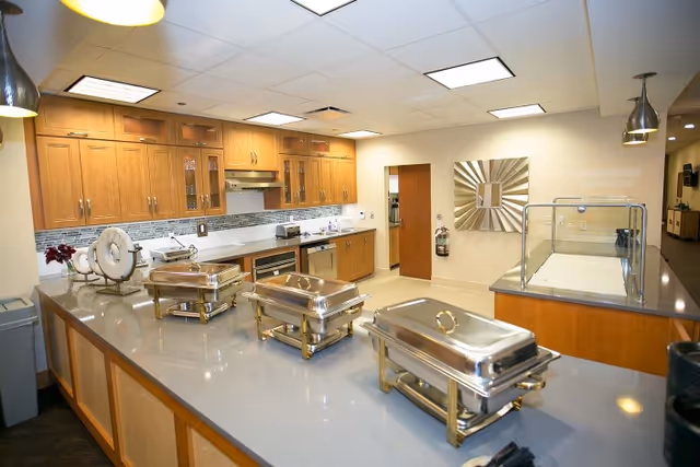 Communal serving kitchen with three stainless steel chafing dishes on a long island, wooden cabinets, and a food service counter.
