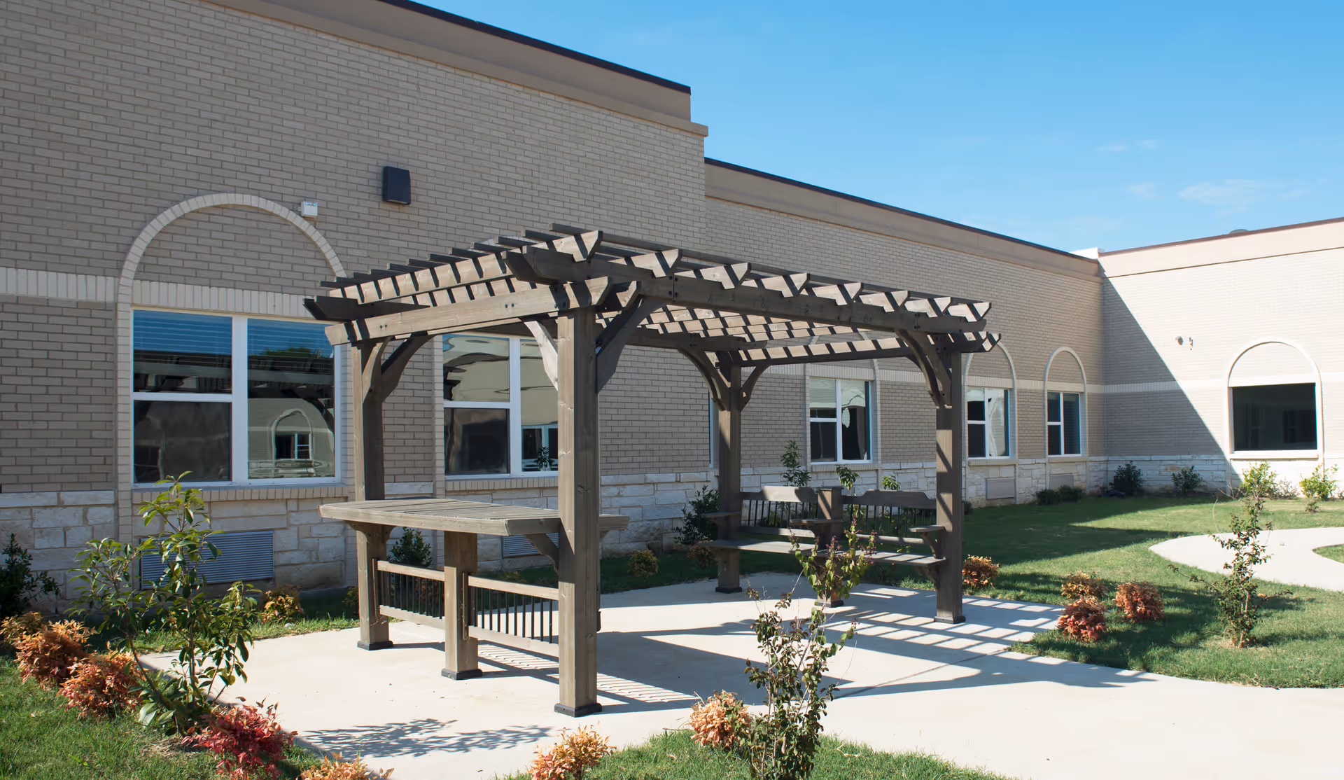 Outdoor seating area with a wooden pergola structure providing partial shade over benches and a table, surrounded by a grassy courtyard with small plants and shrubs, adjacent to a beige brick building with multiple windows.