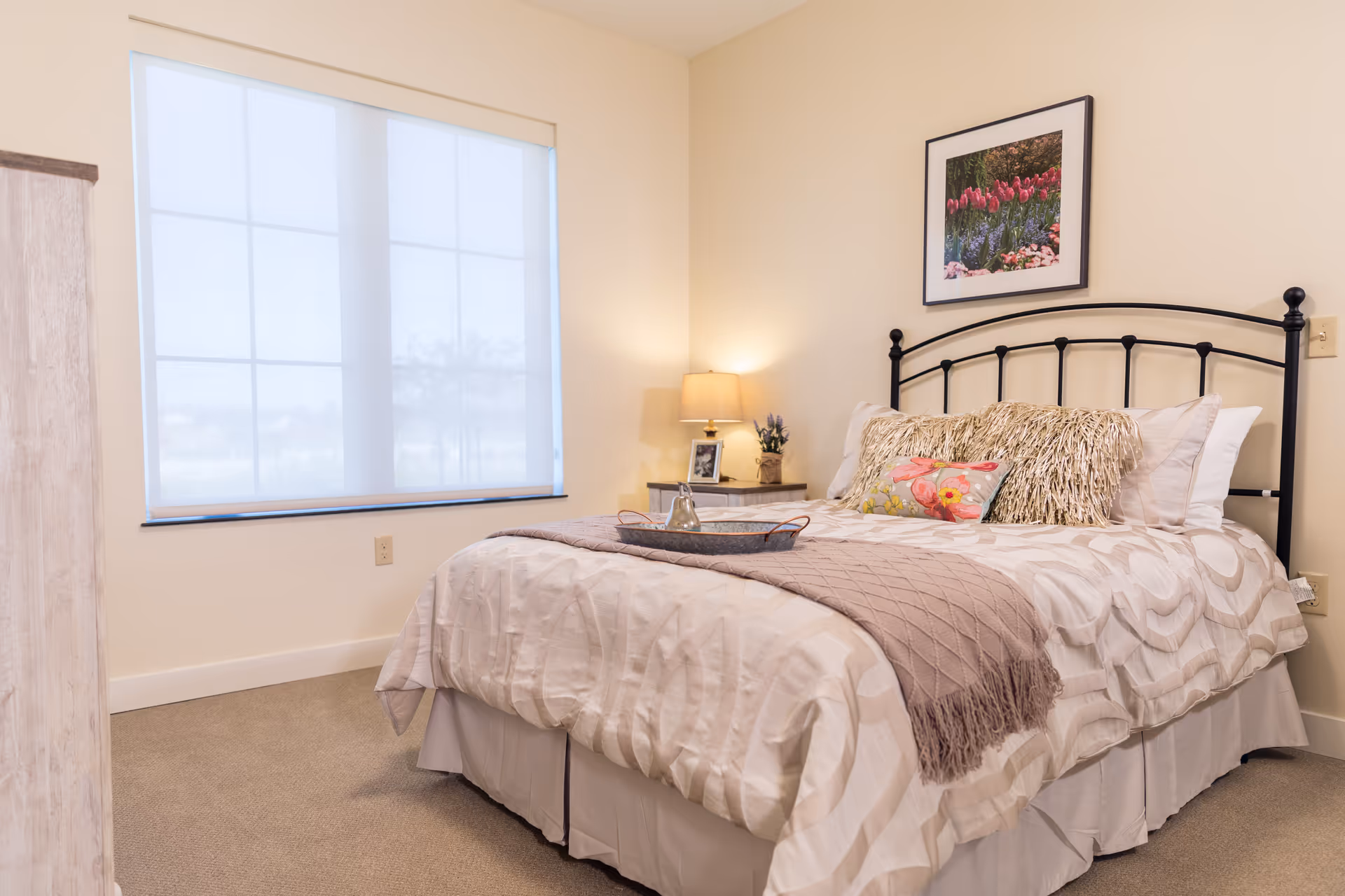 A cozy bedroom with a neatly made bed featuring a white and beige patterned comforter, decorative pillows, and a mauve throw blanket. A black metal headboard is visible, with a framed picture of flowers hanging above it. To the right of the bed is a nightstand with a lamp, a small plant, and a framed photo. A large window with a white roller shade lets in natural light, and beige carpet covers the floor.