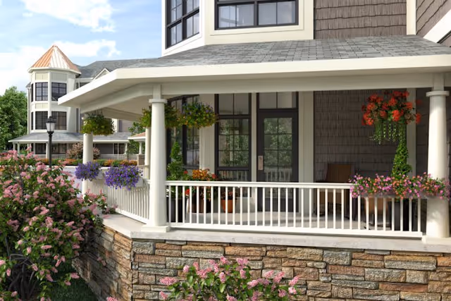 A covered porch area of a senior living facility with white railings and columns, hanging flower baskets, and potted plants. The building has stone and shingle exterior walls, large windows, and a turret-like structure in the background. There are flowering bushes and greenery surrounding the porch.