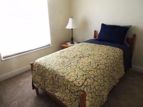 Single bed with a patterned quilt, wooden frame, nightstand, lamp, and window in a small bedroom.
