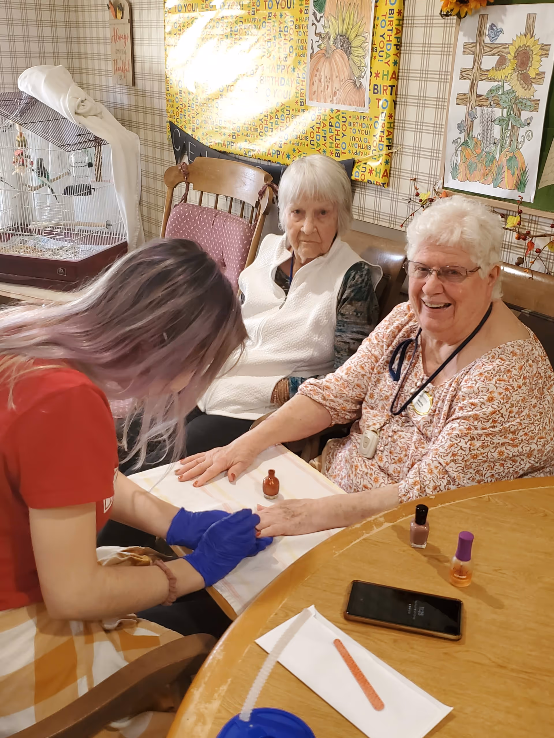 A young woman wearing blue gloves is painting the nails of an elderly woman who is smiling. Another elderly woman is sitting beside her, watching. They are seated at a wooden table with nail polish bottles and a smartphone on it. The background shows a birdcage, chairs, and colorful wall decorations.