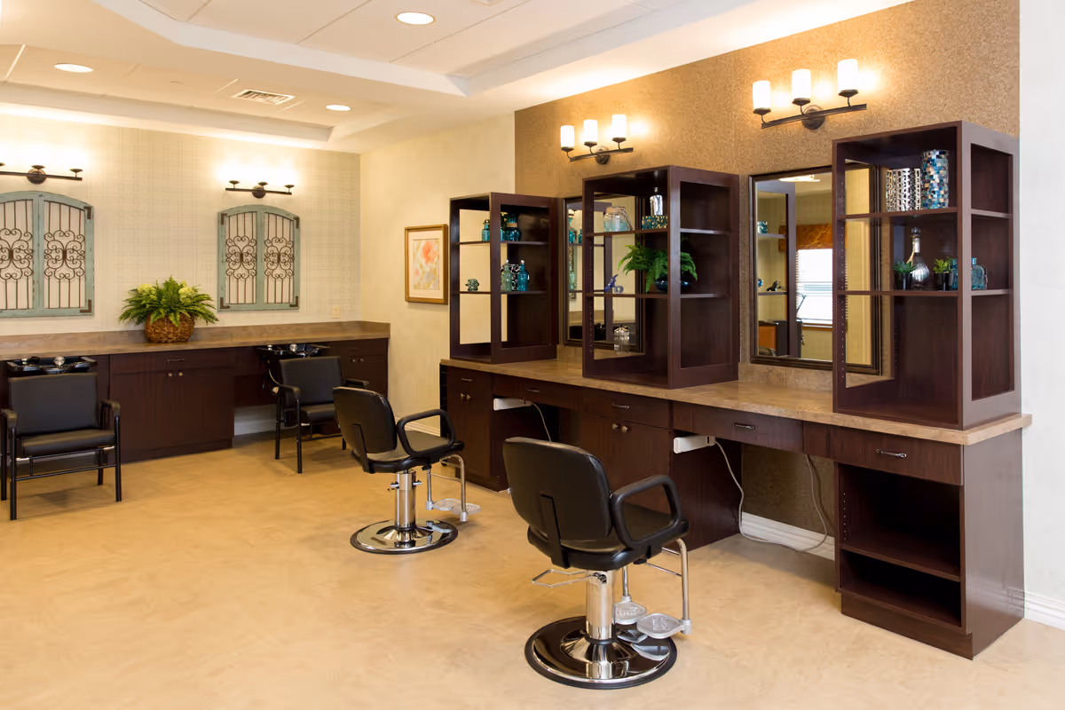 Interior view of a salon area in a retirement community featuring two black salon chairs in front of a long counter with mirrors and shelving units holding decorative items. In the background, there are two black chairs and a countertop with a plant and decorative wall art above it. The room has warm lighting and a beige floor.