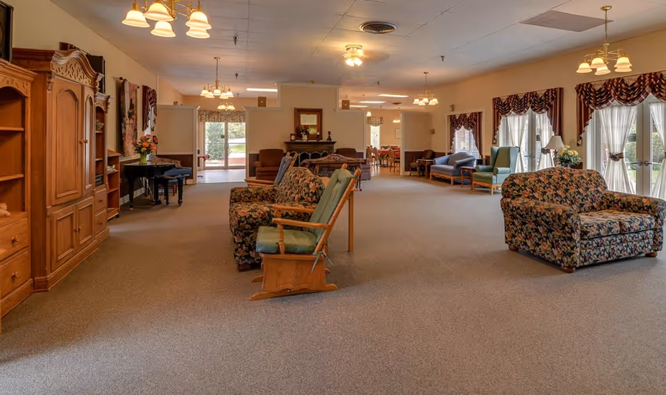 A spacious and well-lit common area in a senior living facility with floral patterned couches, wooden furniture including a rocking chair and a piano, large windows with sheer curtains and valances, and multiple ceiling light fixtures. The room has carpeted flooring and a warm, inviting atmosphere.
