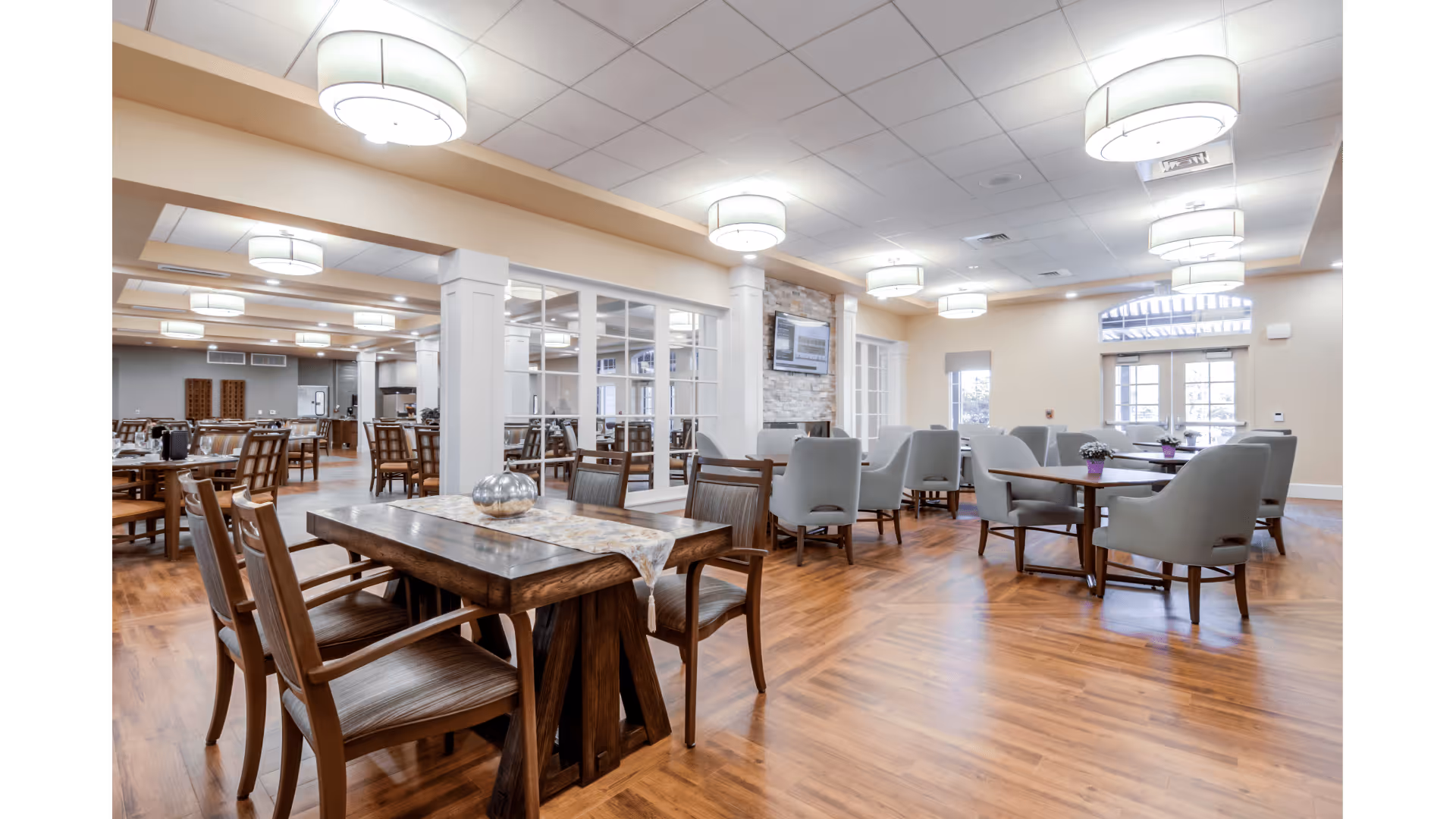 A spacious and well-lit dining area with wooden floors, multiple wooden tables and chairs, and comfortable upholstered armchairs. The room features modern ceiling lights, large windows allowing natural light, and a stone accent wall with a mounted TV. The space is clean and inviting, designed for communal dining and socializing.