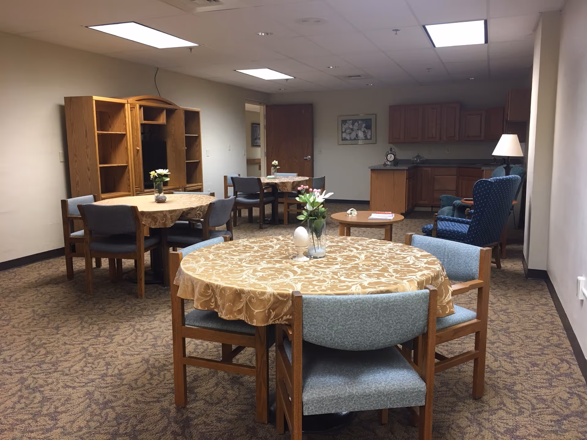 An empty senior living dining area with round tables, chairs, and a kitchenette in back.