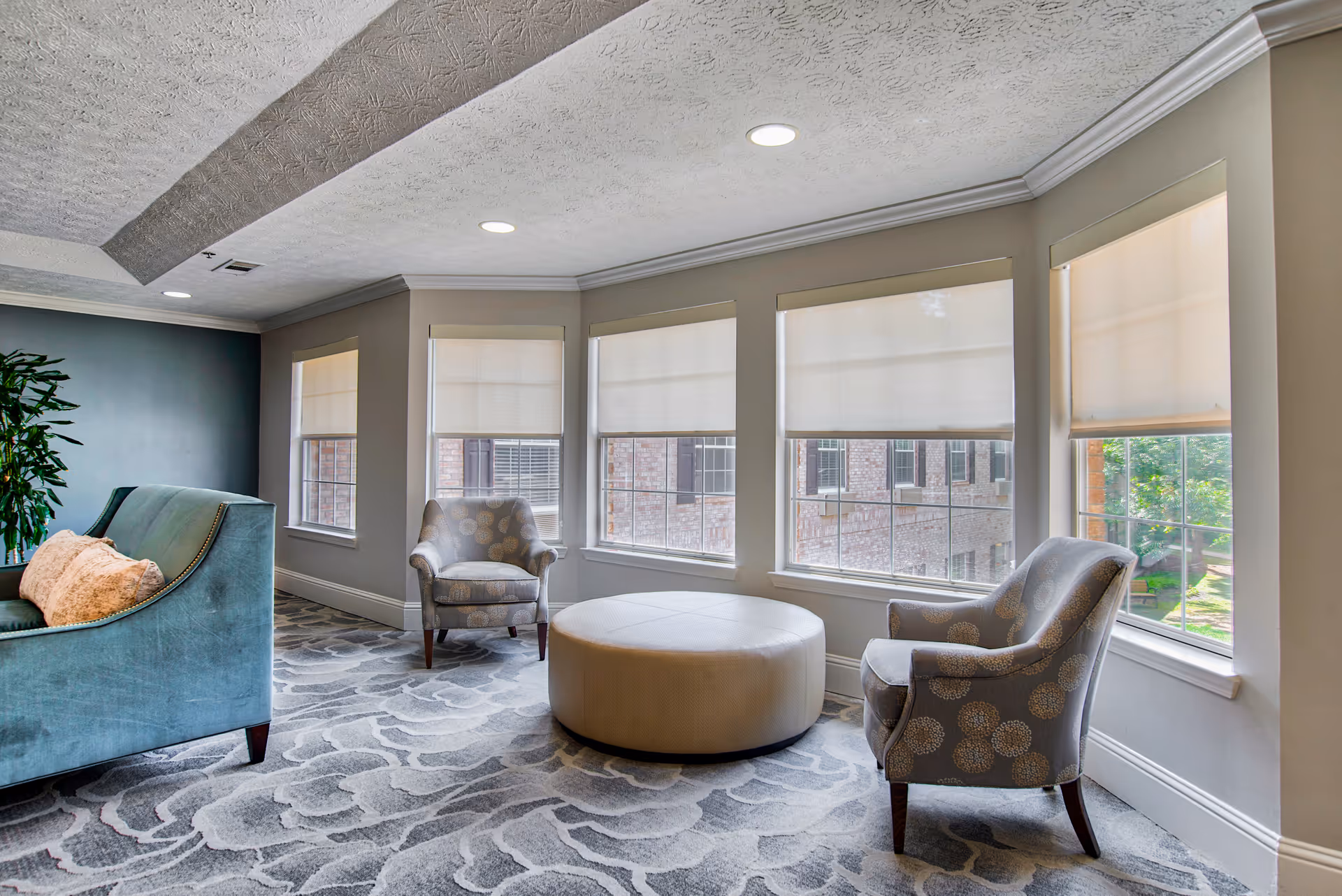 A bright and cozy sitting area with large windows covered by white roller shades. The room features two patterned armchairs, a large round beige ottoman in the center, and a teal sofa with a peach pillow. The floor is carpeted with a floral pattern, and there is a green plant in the corner.