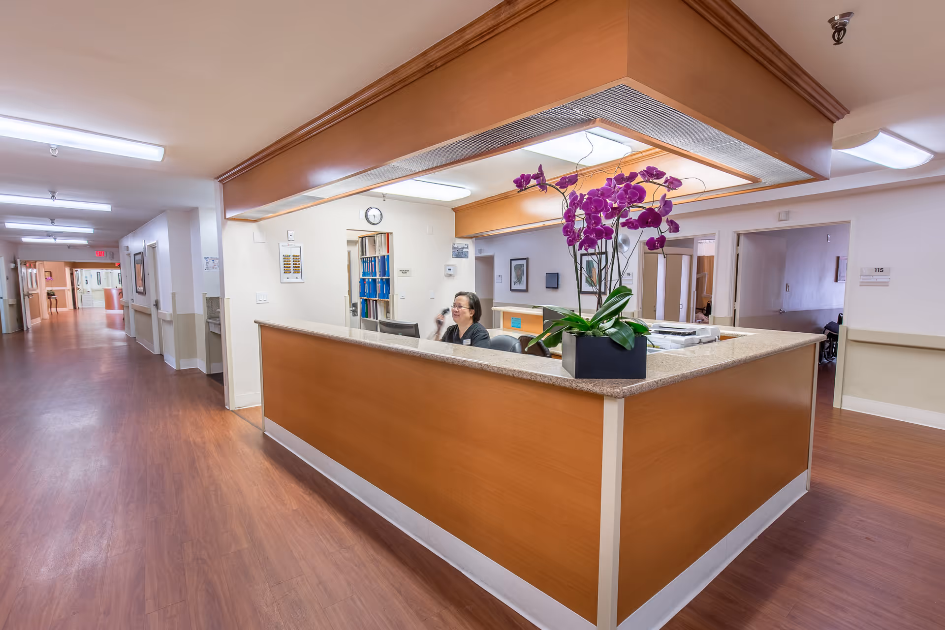 Reception desk area inside a senior living facility with a staff member sitting behind the desk on the phone. The desk has a wooden finish with a granite countertop and a purple orchid plant on it. The hallway has wooden flooring and white walls with several doors leading to rooms.