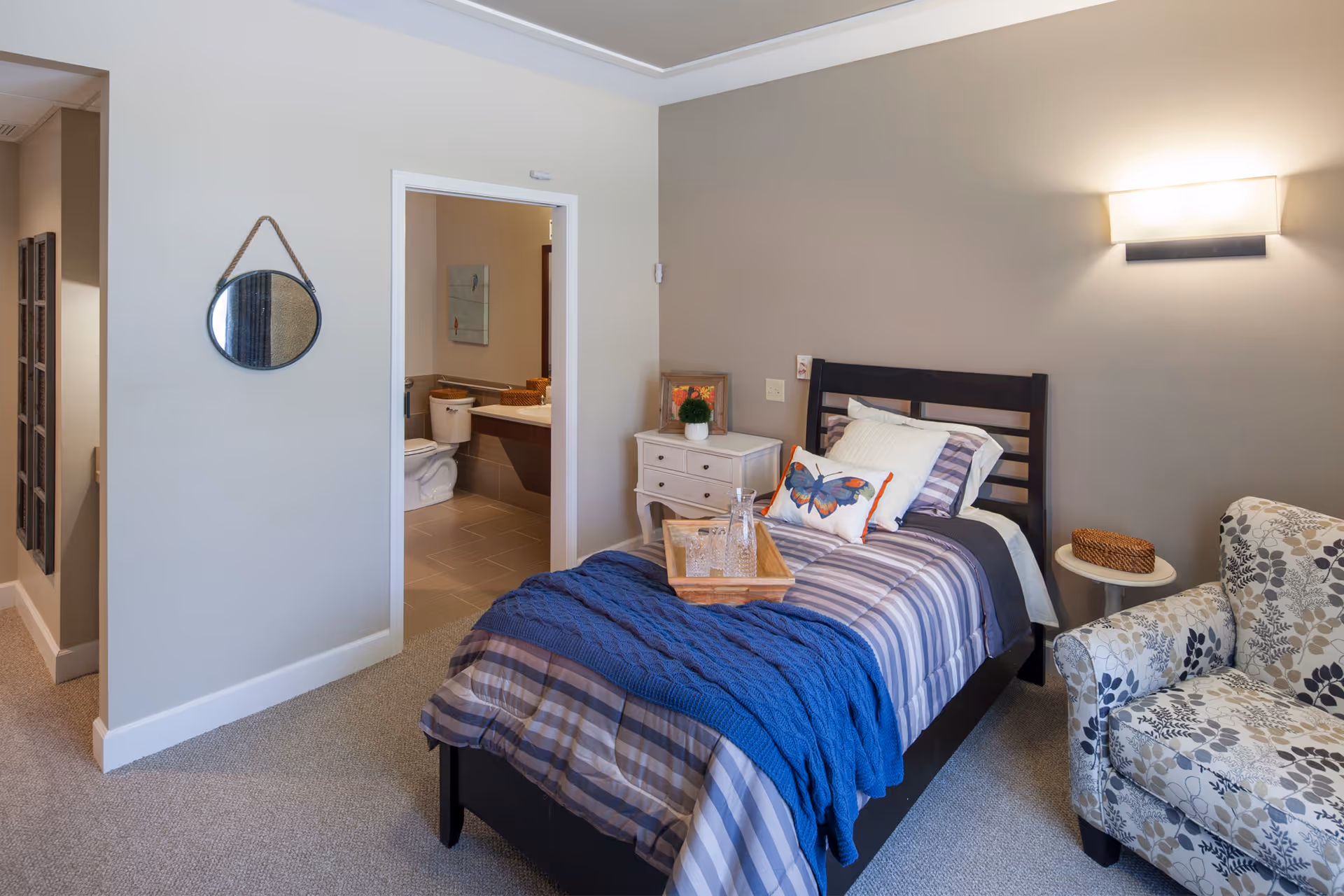 A cozy bedroom with a single bed covered in striped bedding and a blue throw blanket. There is a butterfly-themed pillow on the bed and a wooden tray with a glass pitcher and glasses. Next to the bed is a small white nightstand with a potted plant and a framed picture. A floral patterned armchair and a small round side table with a wicker basket are also visible. The room has beige walls and carpeted floor. A doorway leads to a bathroom with a toilet and sink visible. A round mirror hangs on the wall near the doorway.