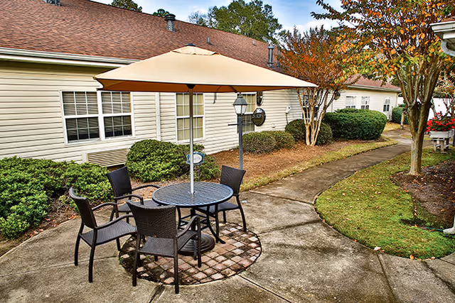 Outdoor patio area with a round table and four chairs under a large beige umbrella. The patio is surrounded by a concrete walkway, bushes, trees, and the exterior walls of a single-story building with windows.